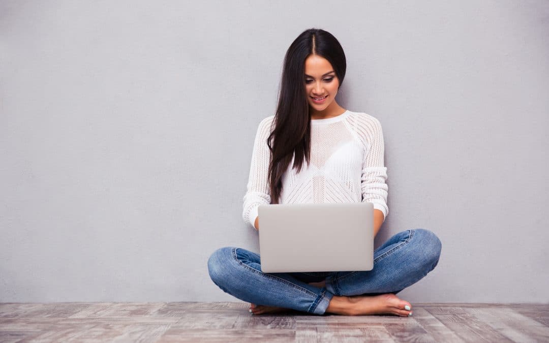 Portrait of a happy casual woman sitting on the floor with laptop on gray background