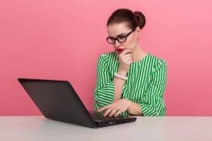 Businesswoman with hair bun in striped blouse and eyeglasses looks seriously at laptop computer isolated on pink background.