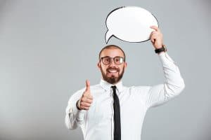 Portrait of a smiling businessman holding empty speech bubble above his head and showing thumbs up isolated over gray background