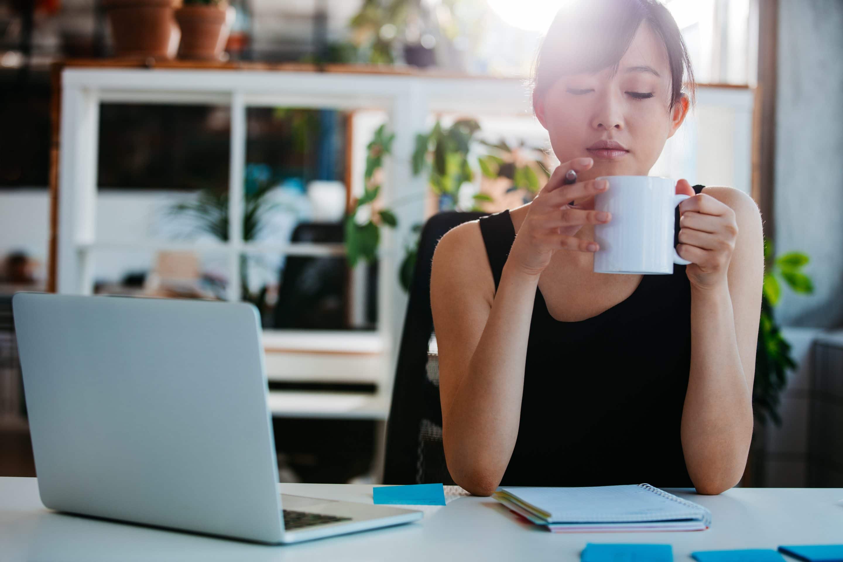 Portrait of relaxed young woman sitting at her desk and having coffee. Asian business woman taking coffee break in office.