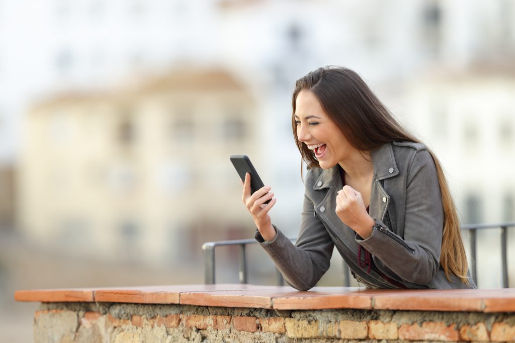 Woman is excited looking at iPhone while outdoors. This is to show that people will be excited to find what they are looking for on search engines if your business shows up first on Google.