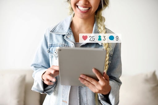 A smiling women holds a tablet with the likes she is getting showing above it. Social media marketing