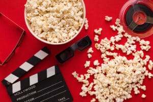 Movie background. Clapperboard, film reel, popcorn and 3D glasses on red backdrop, top view