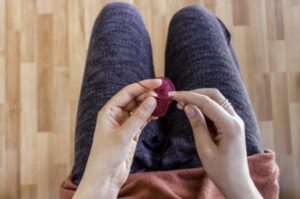 A photo from above of someone knitting with purple string in their lap. Just their hands, legs and string are in the photo. 